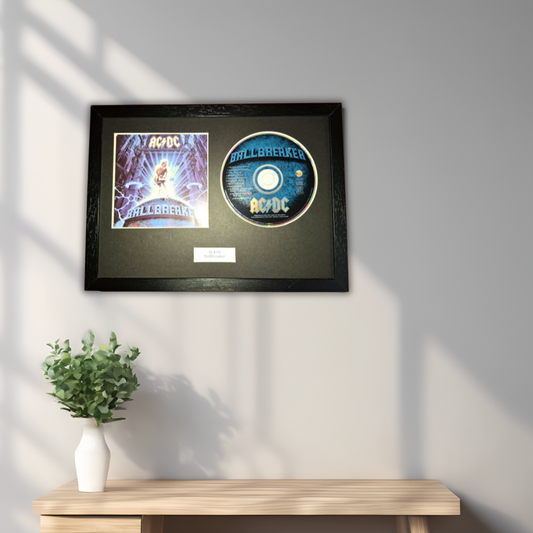 A framed AC/DC 'Ballbreaker' collectible display featuring the album cover and a printed CD, mounted on a dark background and hung on a light gray wall above a wooden console table with a small potted plant.