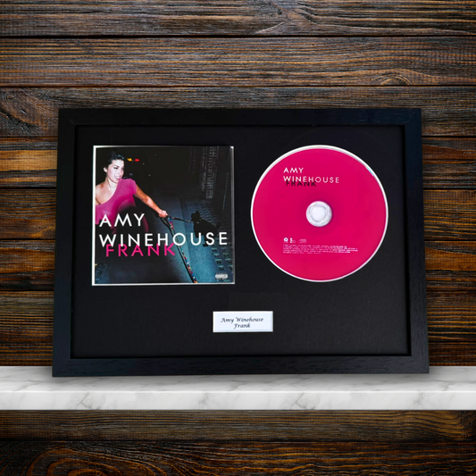 A framed Amy Winehouse 'Frank' CD display featuring the album cover, a pink CD, and a small name plaque inside a black frame, set against a dark wood background.