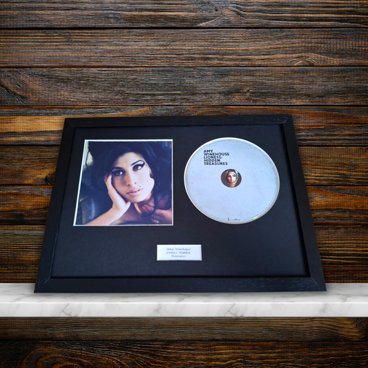 A framed music memorabilia display featuring a black CD case with a printed portrait, a silver CD, and a small white plaque, set in a black frame against a dark wood background.