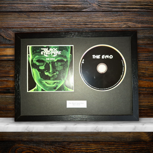 A framed black album print and CD set for the band 'The Blackened', featuring a green-toned square album cover and a black CD with white text, mounted in a black frame against a dark wood background.