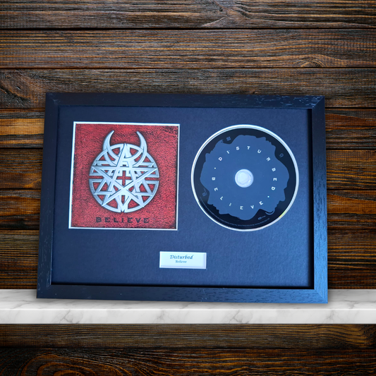 A framed metal CD single for the band 'Believe' by 'Disturbed' on a dark wood background. The black frame contains a red square insert with a silver pentagram logo and the word 'BELIEVE', alongside a black CD with a white border