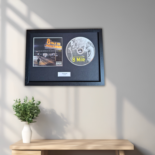 A framed 8 Mile music memorabilia display hanging on a light gray wall above a wooden console table with a small green plant in a white vase. The black frame contains a printed album cover, a silver-colored 8 Mile disc, and a small white plaque.