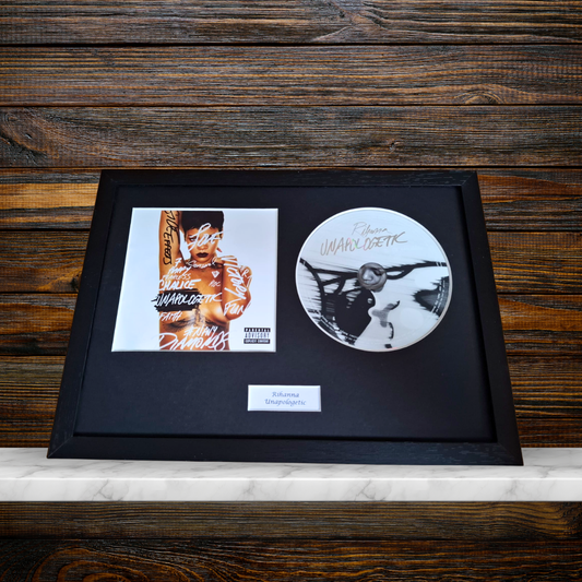A framed music memorabilia display on a white marble surface against a dark wood background, featuring a black album cover print, a circular white vinyl record, and a small plaque.
