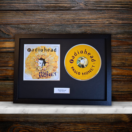 A framed Radiohead 'Pablo Honey' collectible display featuring the yellow CD and the album cover inside a black frame, resting on a white marble surface against a dark wood background.