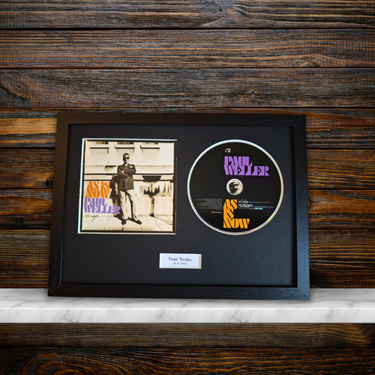 A framed music memorabilia display featuring a vintage CD single of Paul Weller'As Is Now' alongside the original album cover, all mounted in a black frame on a dark wood background.