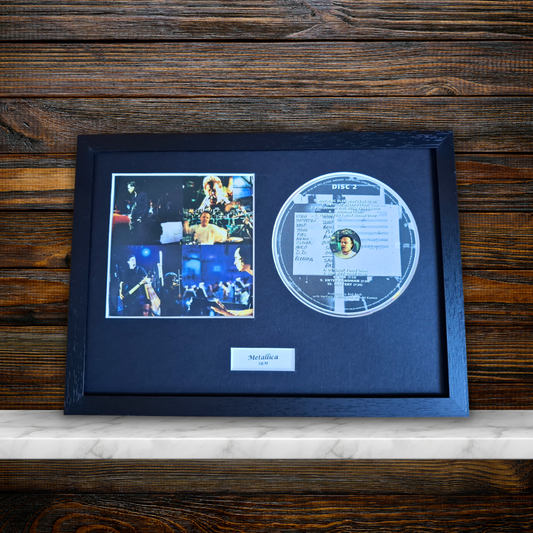 A framed Metallica 'S&M' display featuring a clear acrylic disc, a photo collage, and a nameplate inside a black frame, set against a dark wood background.