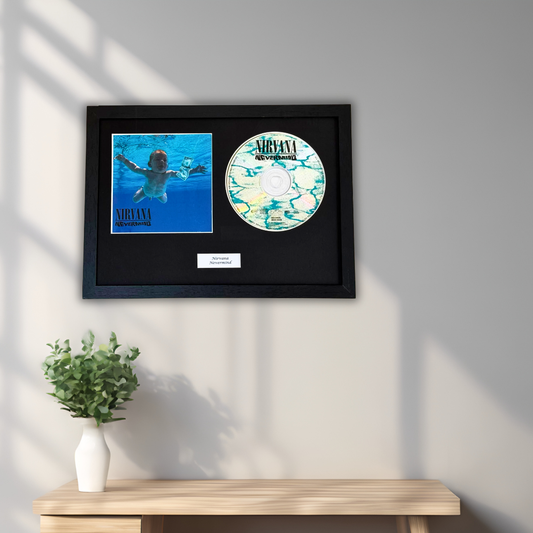 A framed Nirvana memorabilia display hanging on a light gray wall above a wooden console table with a small potted green plant. The black frame contains a blue Nirvana album cover and a circular marbled CD with the Nirvana logo.