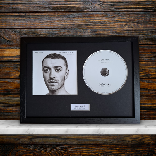 A framed music memorabilia piece featuring a black-and-white portrait print of Sam Smith and a silver-colored CD inside a black frame, mounted on a black mat against a dark wood plank wall with a white marble shelf visible below.
