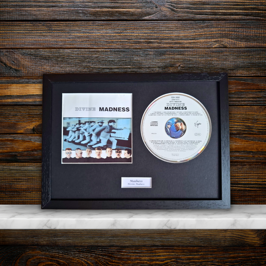 A framed Divine Madness CD album display featuring the original CD, printed cover art, and a small plaque on a black mat inside a dark wood frame, set against a dark wood panel background.