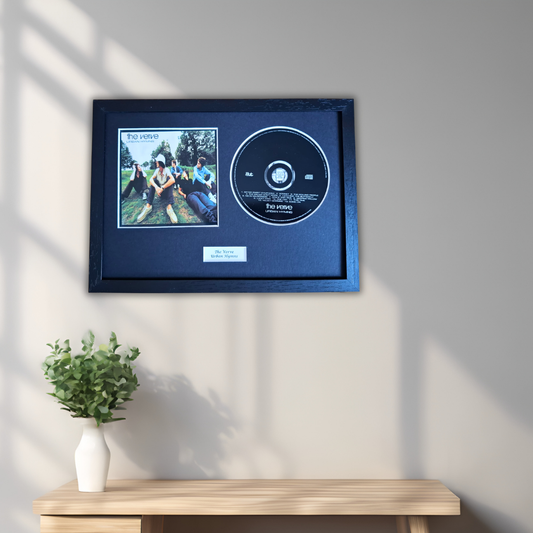 A framed music memorabilia display hanging on a light gray wall above a wooden console table. The frame contains a small square album cover photo and a black CD mounted on a dark navy backing, with a small white plaque below.