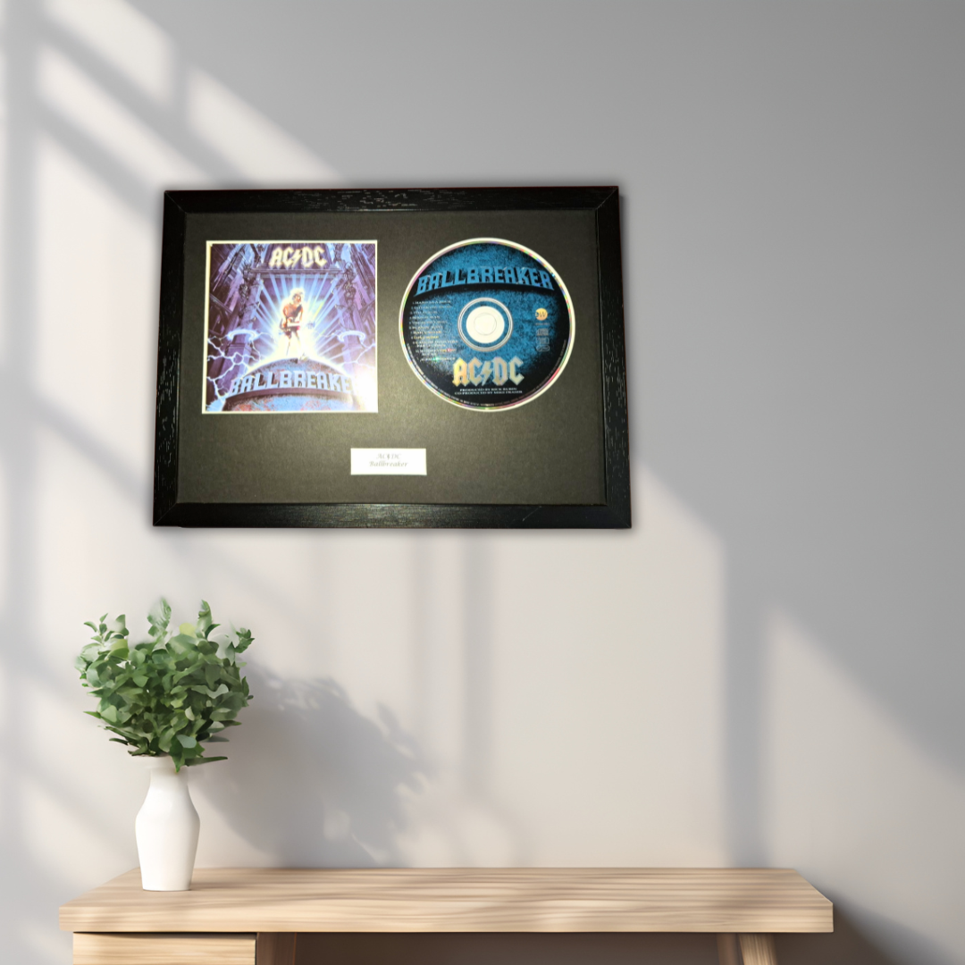 A framed AC/DC 'Ballbreaker' collectible display featuring the album cover and a printed CD, mounted on a dark background and hung on a light gray wall above a wooden console table with a small potted plant.