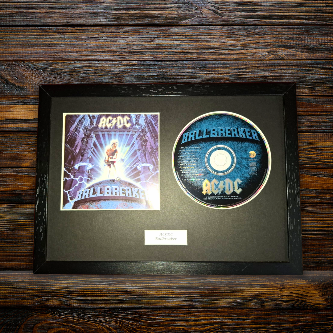 A framed AC/DC 'Ballbreaker' collectible display featuring the original printed album cover and a printed CD on a black mat, set against a dark wood background.