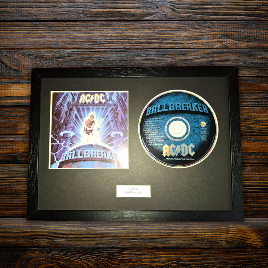 A framed AC/DC 'Ballbreaker' collectible display featuring the original printed album cover and a printed CD on a black mat, set against a dark wood background.