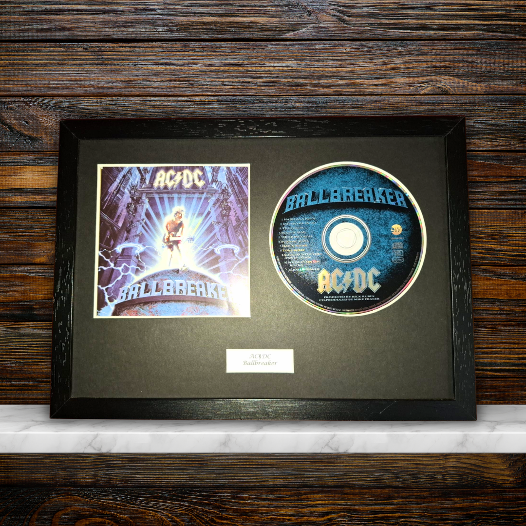 A framed AC/DC 'Ballbreaker' collectible display featuring the original album cover print and a printed CD on a black mat, set against a dark wood background.