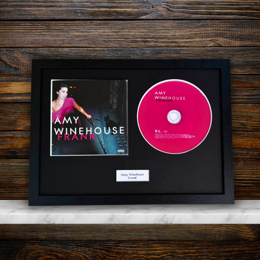 A framed Amy Winehouse 'Frank' CD display featuring the album cover, a pink CD, and a small name plaque inside a black frame, set against a dark wood background.
