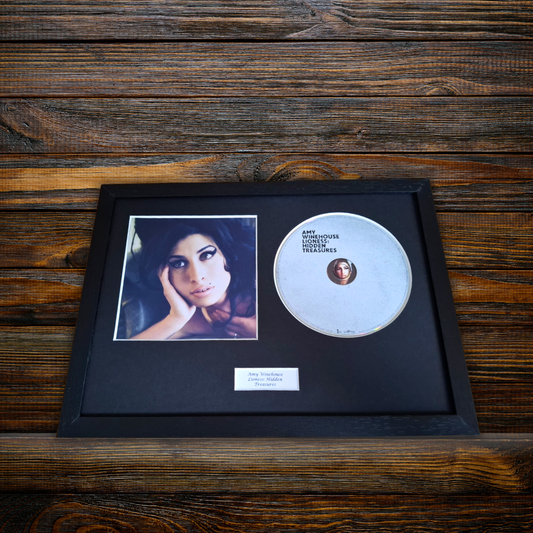 A framed Amy Winehouse 'Lioness: Hidden Treasure" CD featuring a black frame with a black mat, a printed portrait of Amy Winehouse, the silver CD, and a small white plaque, displayed on a dark wood background.