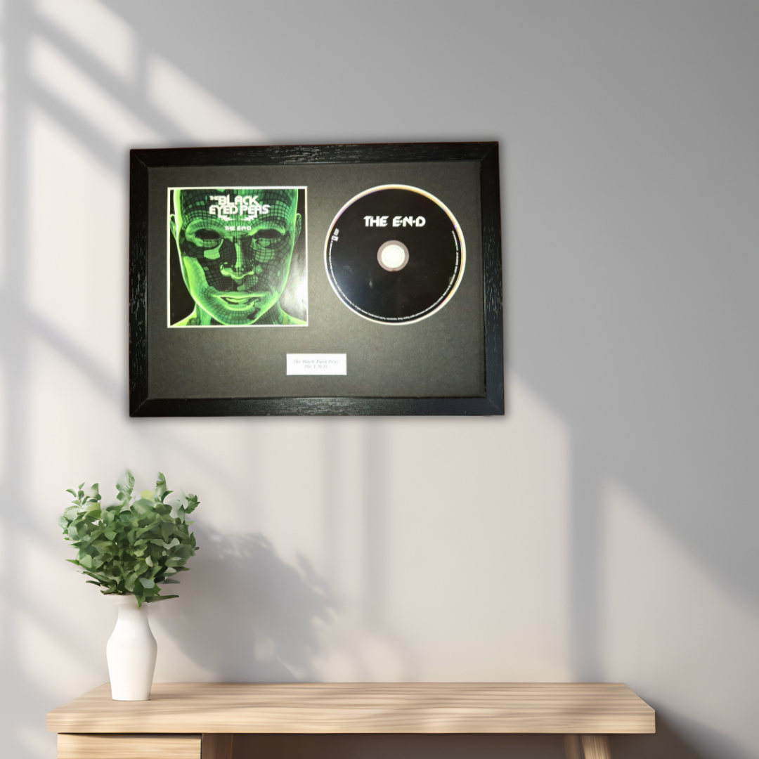 A framed black record plaque featuring a green-toned album cover and a black CD disc on a dark mat, mounted on a light gray wall above a wooden console table with a small white vase of greenery.