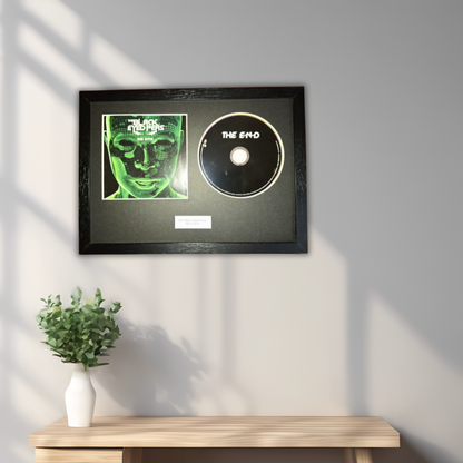 A framed black record plaque featuring a green-toned album cover and a black CD disc on a dark mat, mounted on a light gray wall above a wooden console table with a small white vase of greenery.