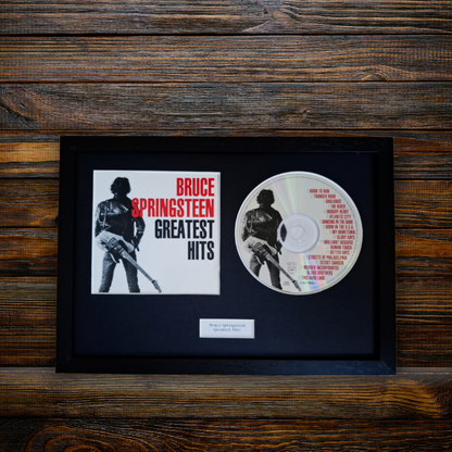 A framed Bruce Springsteen 'Greatest Hits' album display featuring the album cover insert and a printed CD, mounted in a black frame against a dark wood plank background.