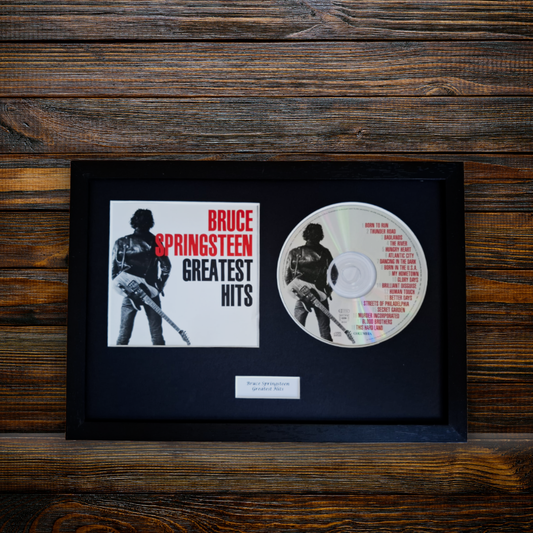 A framed Bruce Springsteen 'Greatest Hits' album display featuring the album cover insert and a printed CD, mounted in a black frame against a dark wood plank background.