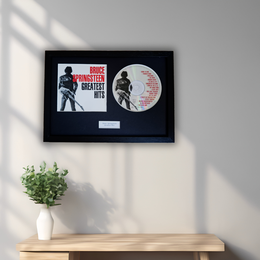 A framed Bruce Springsteen 'Greatest Hits' collectible display mounted on a light gray wall above a wooden console table with a small potted green plant. The display includes a black frame with a black mat, a white insert with red and black text, a circular printed disc, and a small plaque.