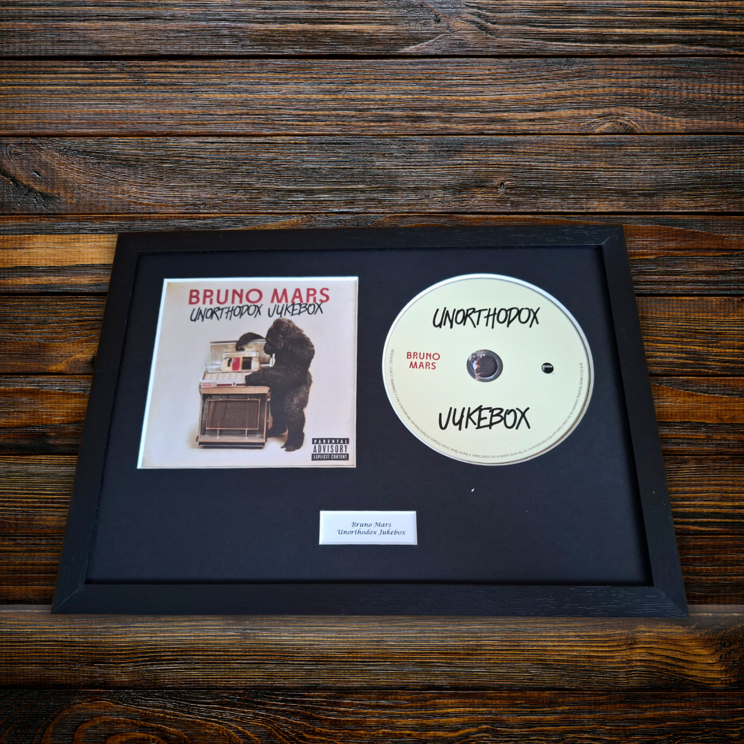 A framed Bruno Mars 'Unothodox Jukebox ' music display featuring the album cover, a custom white CD, and a small plaque on a black mat, set against a dark wood background.