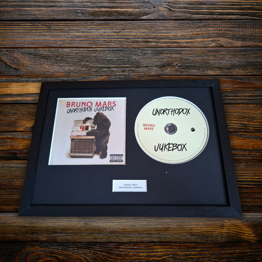 A framed Bruno Mars 'Unothodox Jukebox ' music display featuring the album cover, a custom white CD, and a small plaque on a black mat, set against a dark wood background.