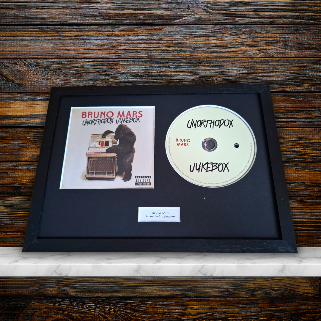 A framed Bruno Mars 'Unorthodox Jukebox' music display featuring the album cover, a custom white CD with black handwritten text, and a small plaque inside a black frame, set against a dark wood background.