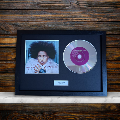 A framed music memorabilia piece featuring a black frame with a portrait photo of Mary Gray, a silver CD with a pink center label, and a small nameplate, displayed against a dark wood background.