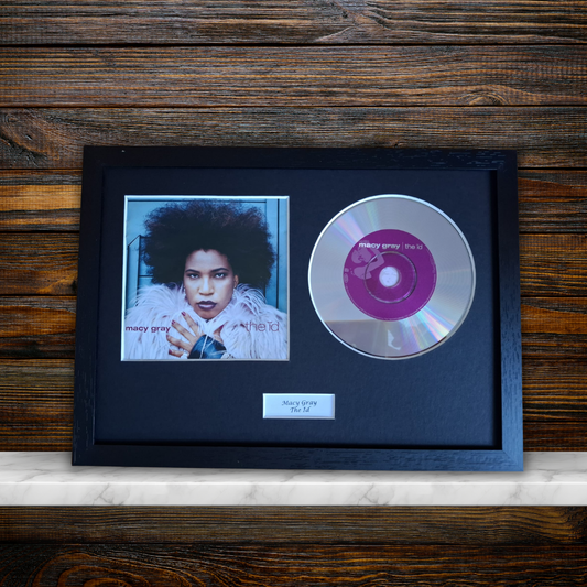 A framed music memorabilia piece featuring a black frame with a portrait photo of Mary Gray, a silver CD with a pink center label, and a small nameplate, displayed against a dark wood background.