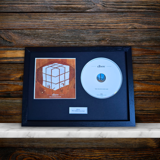 A framed CD single display featuring the album art and disc for 'Elbow' mounted on a dark mat inside a black frame, resting on a light marble surface against a dark wood plank background.