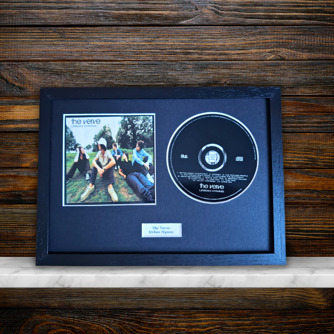 A framed and mounted music memorabilia piece featuring a CD single and a printed album cover for The Verve's 'Urban Hymns' displayed on a light marble surface against a dark wood background.