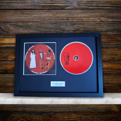 A framed music display featuring a red CD and a printed album cover insert on a dark navy mat, set in a black frame against a rustic dark wood background.