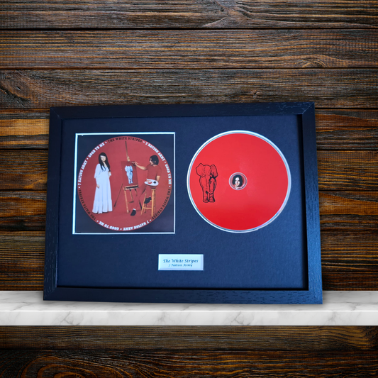 A framed music display featuring a red CD and a printed album cover insert on a dark navy mat, set in a black frame against a rustic dark wood background.