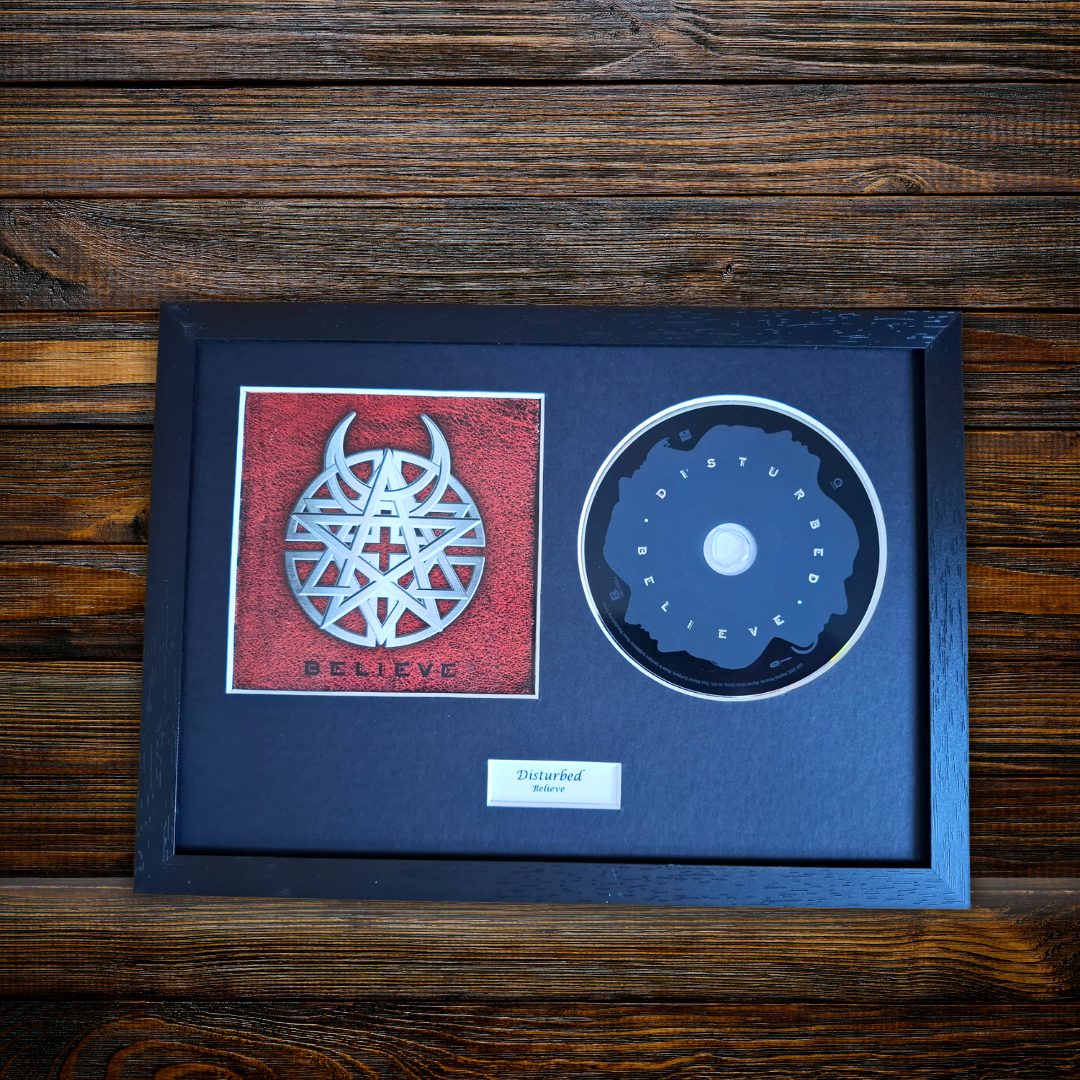 A framed, backlit CD single for the band Disturbed, featuring their 'Believe' album artwork and a backlit 'Disturbed' logo on the CD, all housed in a black frame against a dark wood background.