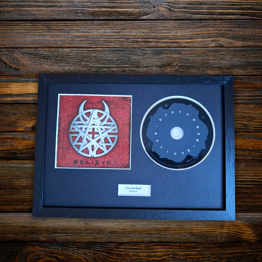 A framed, backlit CD single for the band Disturbed, featuring their 'Believe' album artwork and a backlit 'Disturbed' logo on the CD, all housed in a black frame against a dark wood background.
