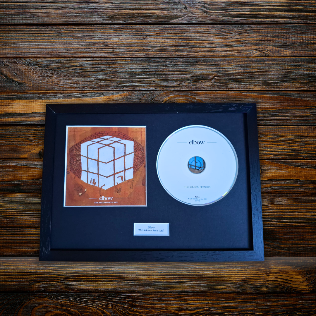 A framed CD single for the band 'elbow' featuring the album 'The Seldiom Seen Kid' on a dark wood background. The black frame contains a black mat, the square album cover on the left, a silver CD on the right, and a small white label at the bottom.