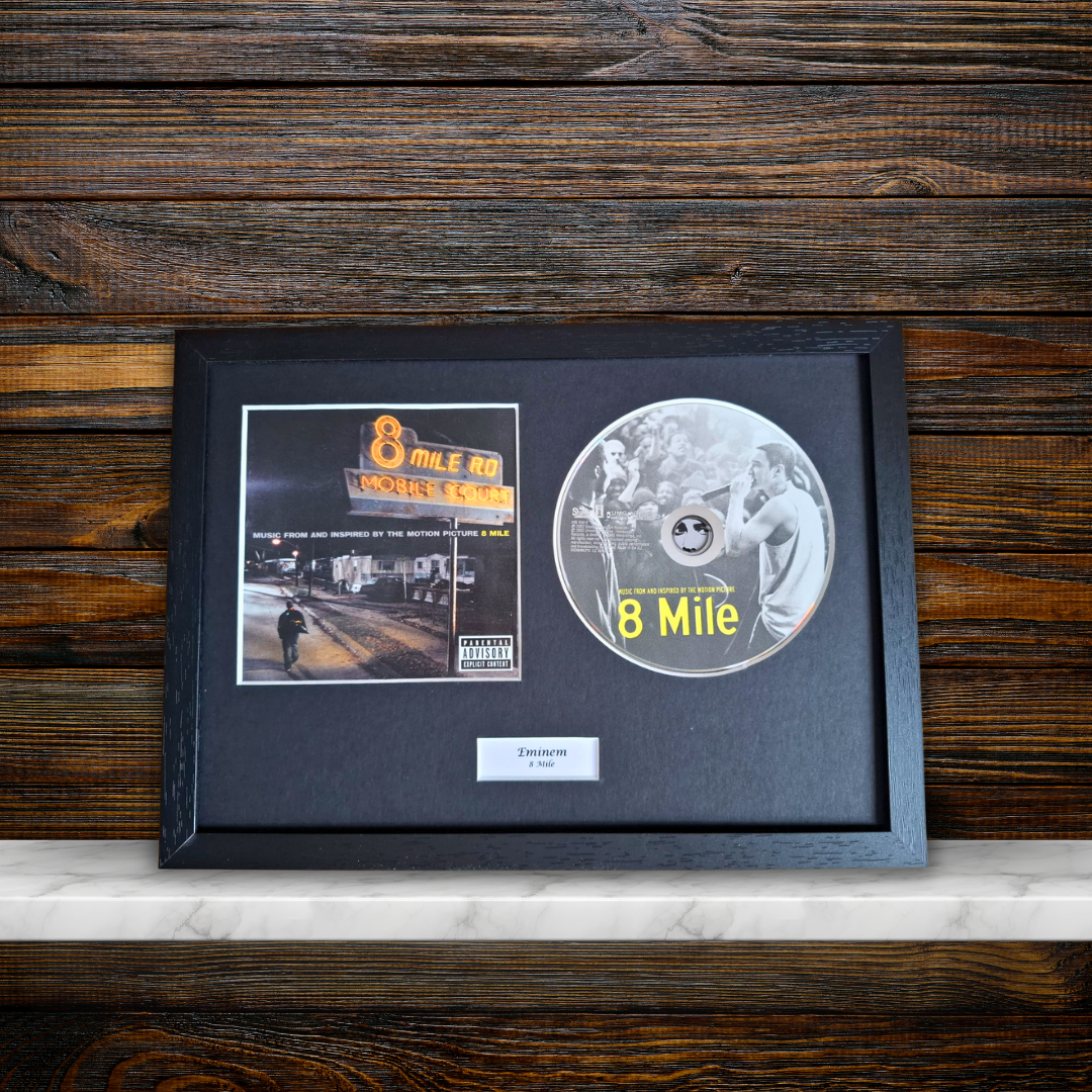 A framed 8 Mile album memorabilia display featuring the original CD, a printed album photo, and a small plaque on a black mat, set against a dark wood plank background.