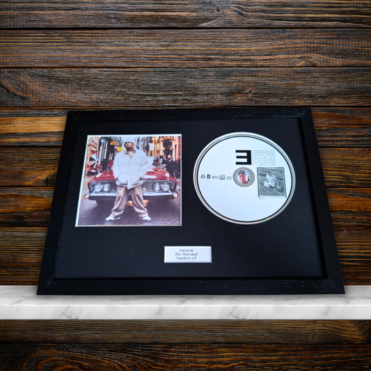 A framed music memorabilia piece featuring a CD and a printed photo of Eminem mounted on a black background with a small plaque, displayed on a marble surface against a dark wood wall.