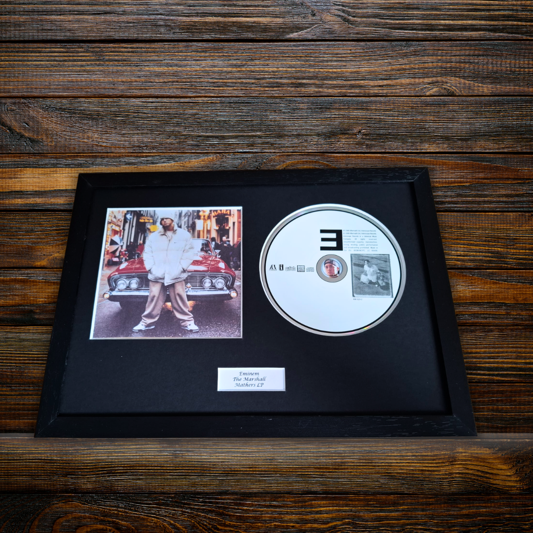 A framed black CD single-player display on a dark wood background. The black frame contains a black mat with a printed photo of a man standing next to a red car, a silver CD with a large black '3' on the front, and a small white label at the bottom.