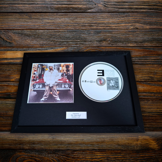 A framed black CD single-player display on a dark wood background. The black frame contains a black mat with a printed photo of a man standing next to a red car, a silver CD with a large black '3' on the front, and a small white label at the bottom.