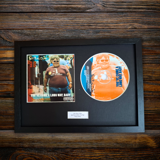 A framed music memorabilia piece featuring a printed album cover and a circular printed disc on a black mat, displayed against a dark wood plank background.