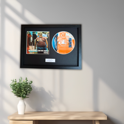 A framed memorabilia display hanging on a light gray wall above a wooden console table with a small potted green plant. The black-framed piece contains a printed photo and a circular orange-and-blue disc.
