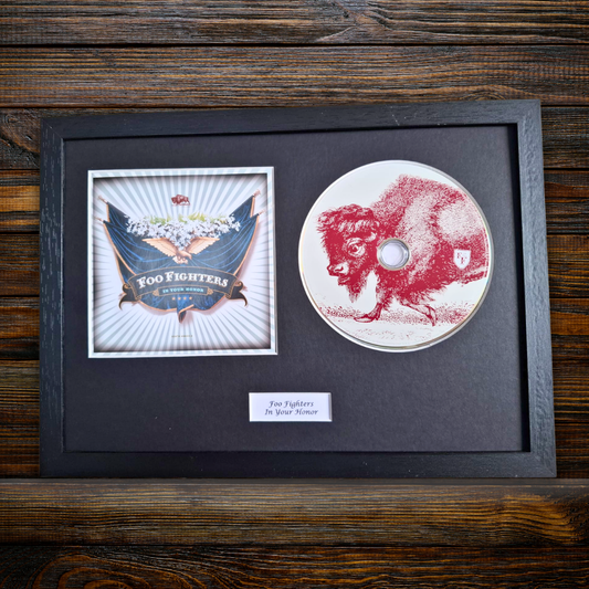 A framed Foo Fighters 'In Your Honor' CD display on a dark wood background. The black-framed display contains the CD, a printed album cover insert, and a small label.