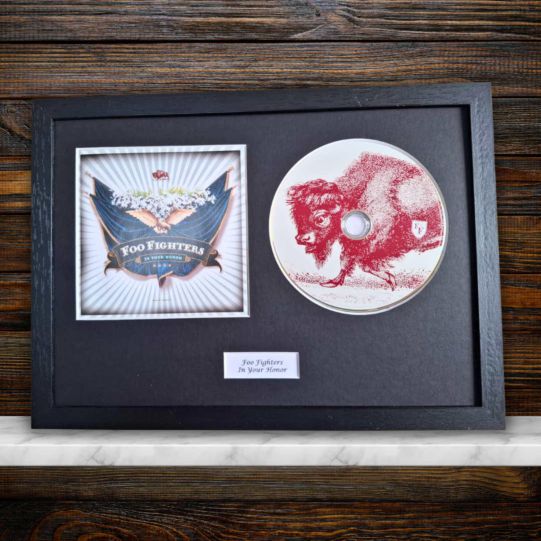 A framed Foo Fighters collectible display featuring the original 'In Your Honor' CD single with a red-buffalo print, alongside the album cover art, all mounted on a black mat inside a black frame with a small label plaque.