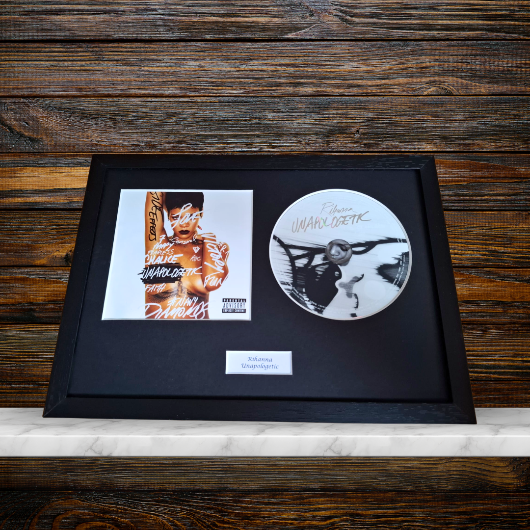 A framed music memorabilia display on a white marble surface against a dark wood background, featuring a black album cover print, a circular white vinyl record, and a small plaque.