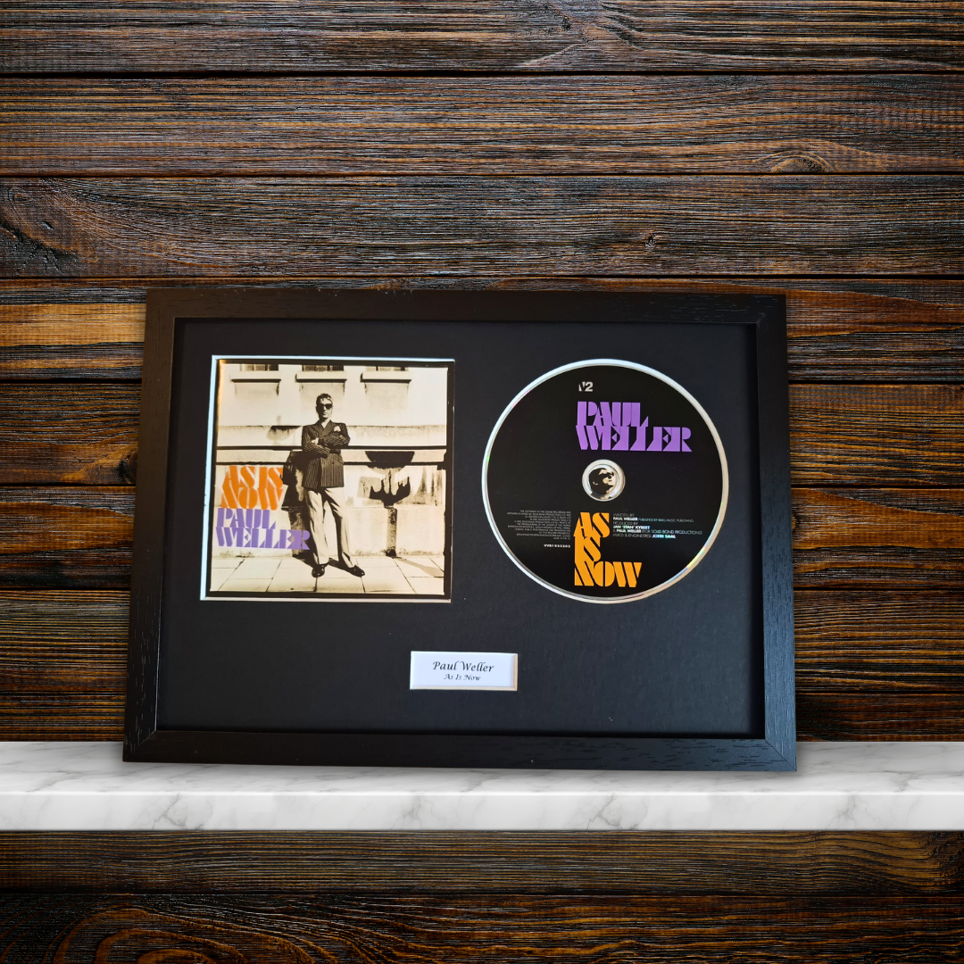 A framed music memorabilia display featuring a vintage CD single of Paul Weller'As Is Now' alongside the original album cover, all mounted in a black frame on a dark wood background.