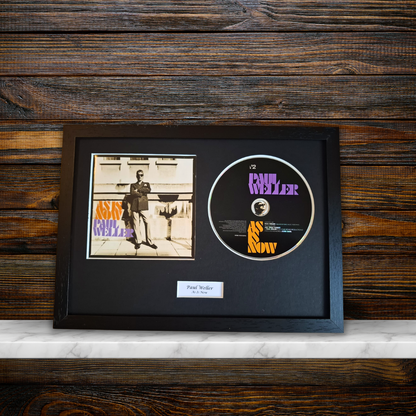 A framed music memorabilia display featuring a vintage CD single of Paul Weller'As Is Now' alongside the original album cover, all mounted in a black frame on a dark wood background.