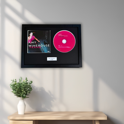 A framed Amy Winehouse 'Frank' CD vinyl record display mounted on a light gray wall above a wooden console table with a small green plant. The display features a black frame with the album cover and a bright pink record sleeve.