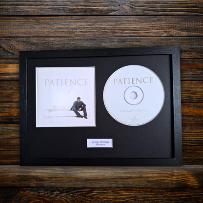 A framed music memorabilia display featuring George Michael's album 'Patience' on a rustic dark wood background. The black frame contains the square album cover, a silver CD, and a small white nameplate reading 'George Michael - Patience'.